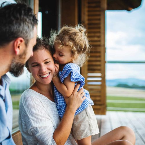 Family,With,Small,Daughter,Sitting,On,Patio,Of,Wooden,Cabin,